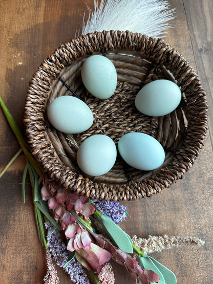 Five light blue eggs in a woven basket on a wooden surface with flowers.