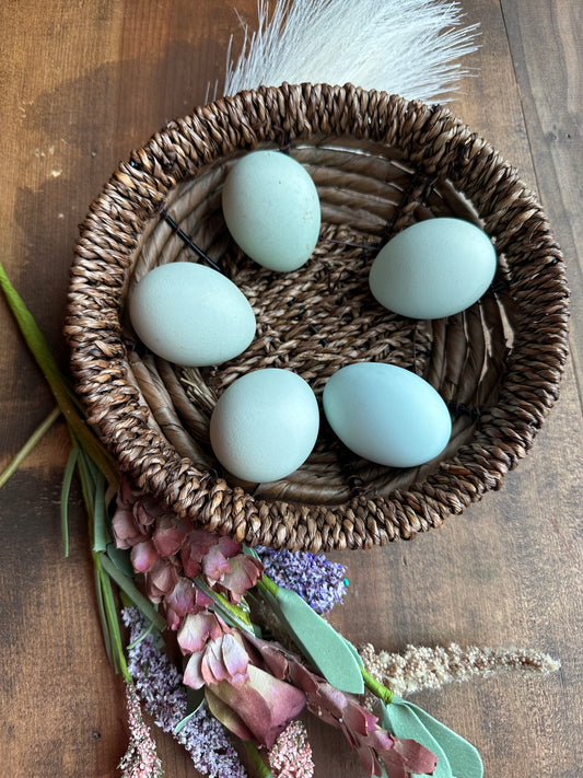 Five light blue eggs in a woven basket on a wooden surface with flowers.